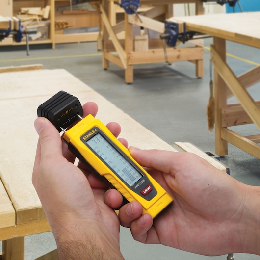 Photo of STANLEY 0-77-030 digital moisture meter held in hands in a woodworking workshop.