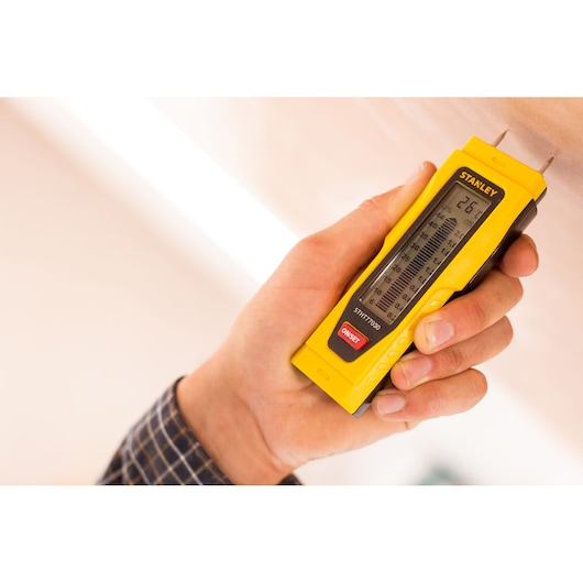 Photo of STANLEY 0-77-030 moisture meter held in hand, yellow body, electronic display, testing a wooden surface.
