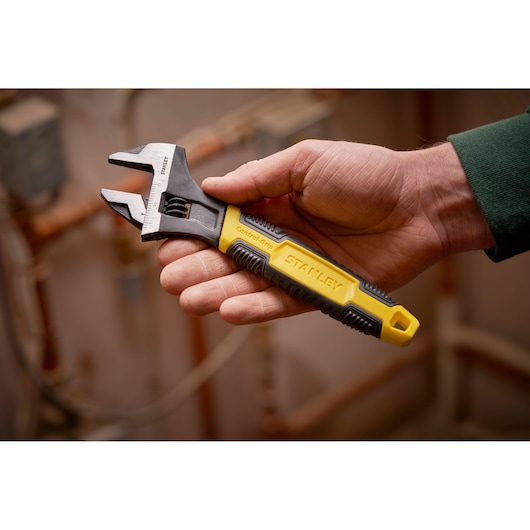 STANLEY 0-90-950 adjustable wrench with yellow and black ControlGrip handle shown in hand, plumbing background.
