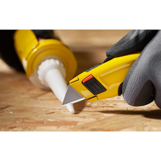 STANLEY 1-11-700 utility knife close-up cutting caulking tube nozzle on wood surface, photo from side angle.
