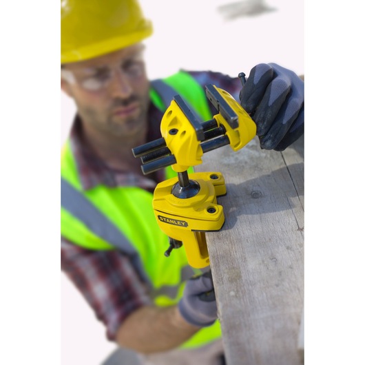 STANLEY 1-83-069 bench vise clamped to wood, side angle, yellow tool, worker in safety gear visible.
