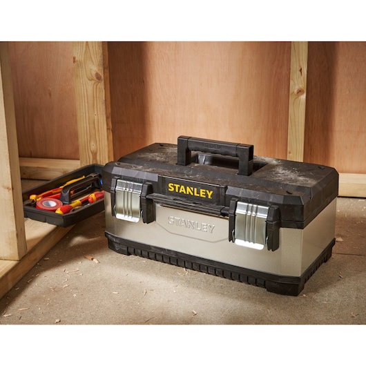 A STANLEY branded metal and plastic toolbox with two latches, placed on a workshop floor. An open tray containing hand tools is visible beside the toolbox.