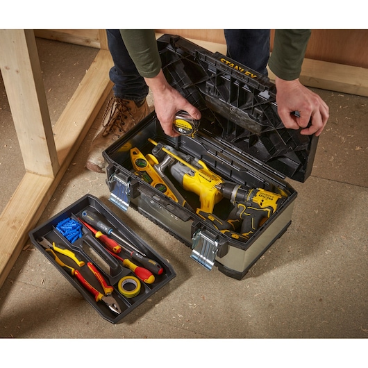 A person opens a STANLEY toolbox filled with various tools such as a drill, measuring tape, level, and hand tools. The removable tray contains additional hand tools and accessories. The toolbox is placed on the floor next to a wooden structure.
