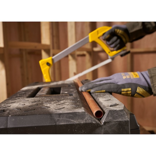 A person wearing work gloves uses a yellow and silver hacksaw to cut a copper pipe on a workbench in a construction setting.
