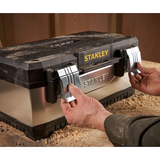 Hands opening metal latches on a STANLEY toolbox resting on a workbench covered in wood shavings.