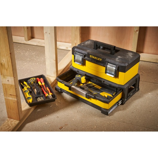 A yellow and black Stanley toolbox with an open drawer showing various hand tools, placed on a construction site floor next to wooden framing.
