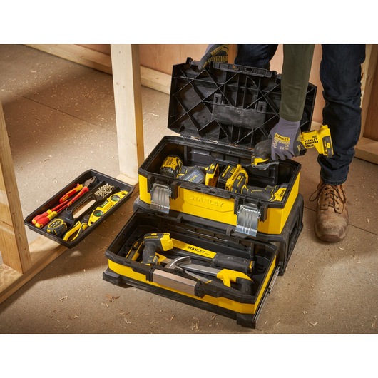 A STANLEY yellow and black toolbox with compartments open and filled with various hand tools and power tools, including drills, screwdrivers, measuring tape, pliers, and a hammer, placed on a construction site floor next to wood framing. A person wearing work gloves is holding a power tool.