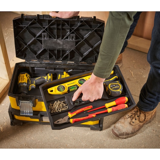 A person opens a STANLEY toolbox, revealing various tools including a level, tape measure, pliers, screwdrivers, and screws on a removable tray. The toolbox is yellow and black, and a power drill is visible inside.