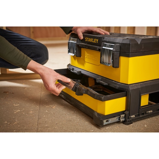 A person opens the bottom drawer of a yellow and black STANLEY toolbox with metal latches, placed on a workshop floor.