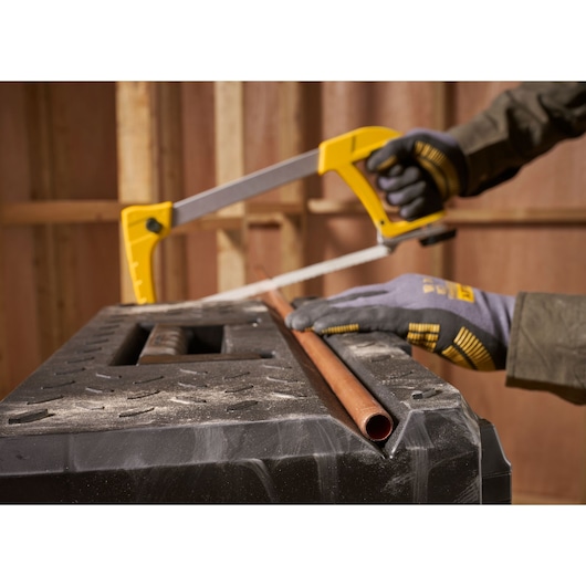A person wearing gloves is using a yellow Stanley hacksaw to cut a copper pipe placed on a black workbench inside a workshop.