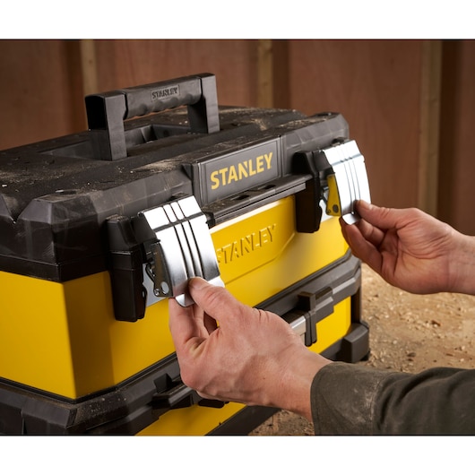 A person closing the metal latches of a yellow and black Stanley toolbox on a dusty work surface.