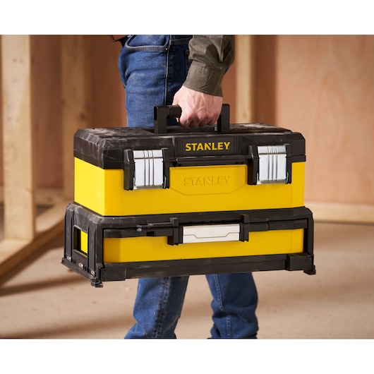 A person is holding a yellow and black STANLEY toolbox with metal latches, standing in an unfinished room with wooden walls.