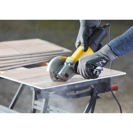 Photo of STANLEY FMEG220K angle grinder cutting ceramic tile on a workbench, close-up side view.