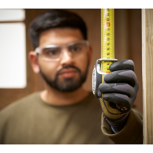 Photo of STANLEY FMHT38214-0 tape measure held vertically by gloved hand, measuring wooden beam indoors.