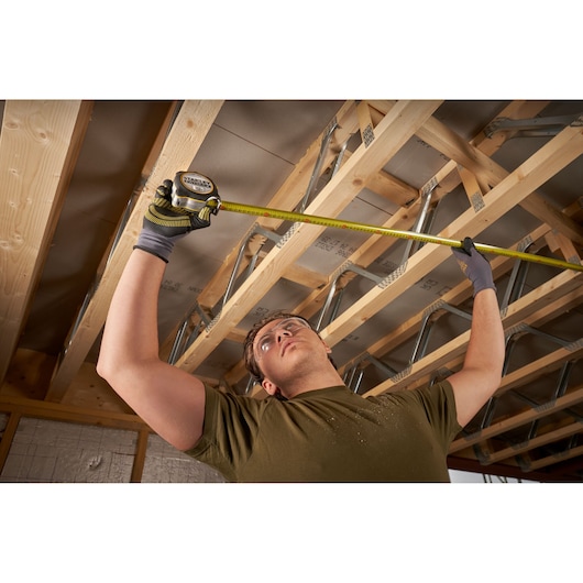 Photo of a worker measuring ceiling joists with a STANLEY FMHT38232-0 tape measure in a construction area.