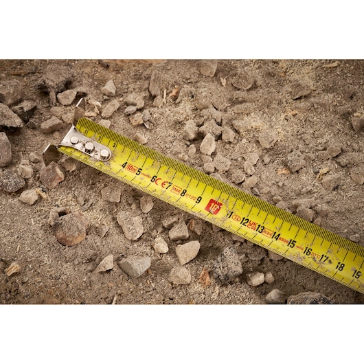 STANLEY FMHT38232-0 tape measure on rocky soil, close-up of yellow measuring tape with black and red markings.