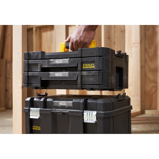 STANLEY FMST1-71969 black portable toolbox with yellow handle being lifted, shown above another toolbox in workshop.