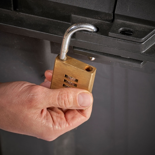 STANLEY FMST17870-1 brass combination padlock held near black storage latch, close-up photo.