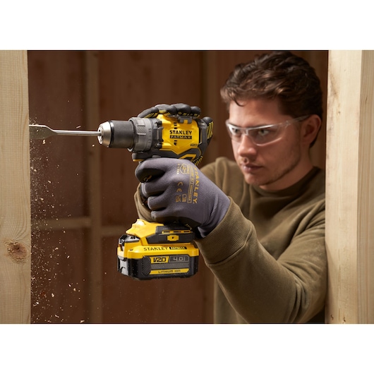 STANLEY SFMCD725B cordless drill in use drilling wood, close-up side view, glove and tool details visible.
