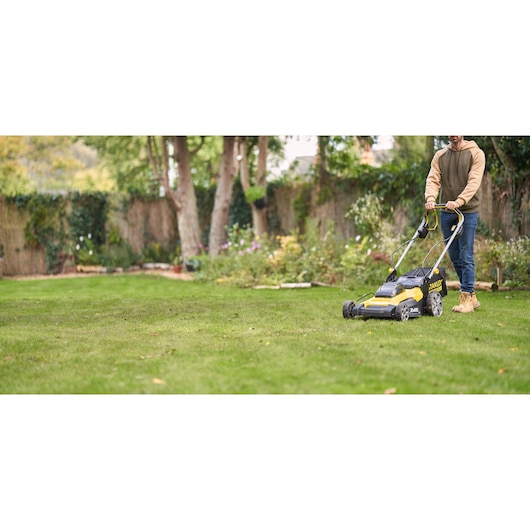Photo of STANLEY SFMCMWS251B cordless lawn mower in use on a green lawn, garden and trees in background.