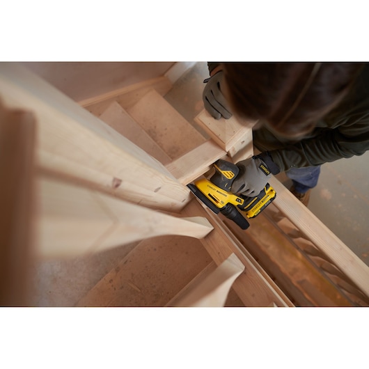 Photo of worker using STANLEY SFMCW221B sander on wooden staircase, wearing gloves, indoor woodworking setting.