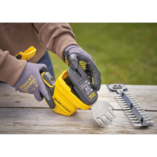 Photo of STANLEY STCSS20B garden shear with battery being inserted, gloved hands, additional blade on table.