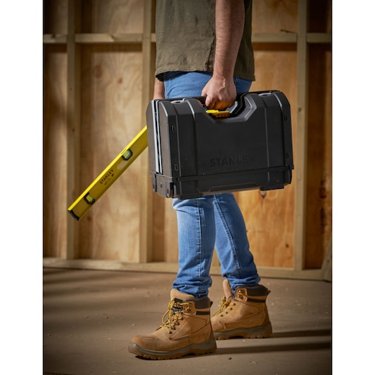 STANLEY STST1-71963 black toolbox carried by person, yellow handle, construction site background, side angle.