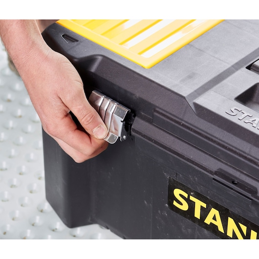 STANLEY STST82976-1 toolbox close-up, hand unlocking metal latch on corner, black and yellow plastic case.
