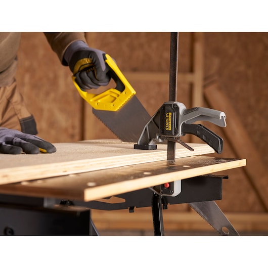 Photo of STANLEY STST83800-1 clamp securing wood on workbench during sawing, woodworking tools visible.